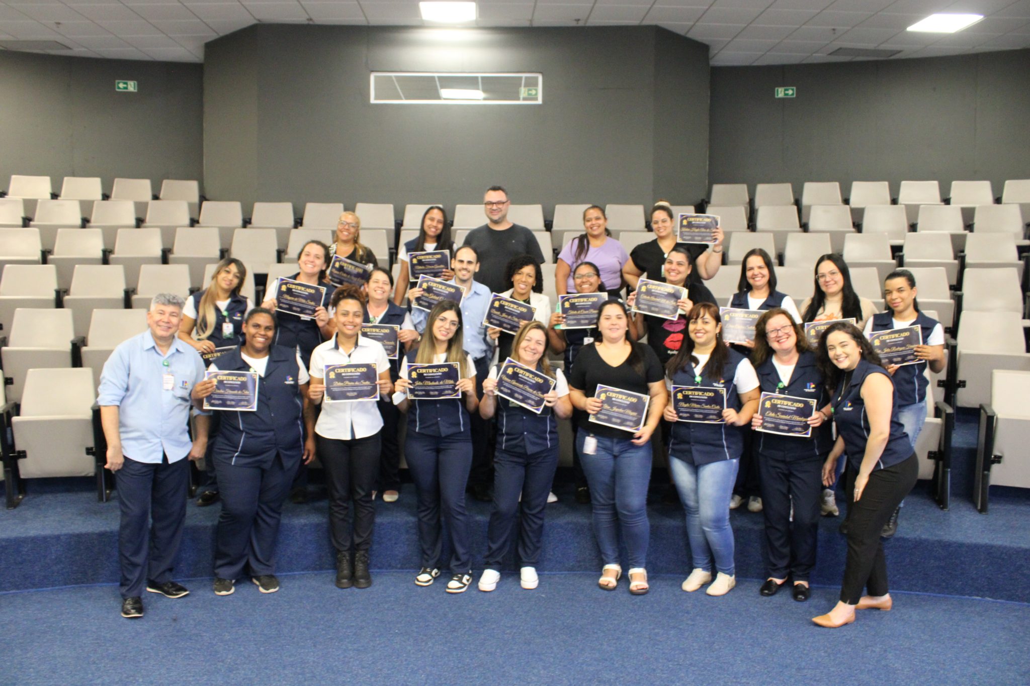 A imagem mostra um grande grupo de pessoas, principalmente mulheres, sentadas em degraus em uma sala de aula. Elas estão segurando certificados e sorrindo para a câmera. Um homem está parado atrás delas. O cenário é uma sala de aula com cadeiras em degraus. A iluminação é boa, e o foco está nas pessoas na frente. Elas parecem estar celebrando uma conquista. A foto sugere uma cerimônia de formatura ou premiação. O ambiente é formal e alegre.