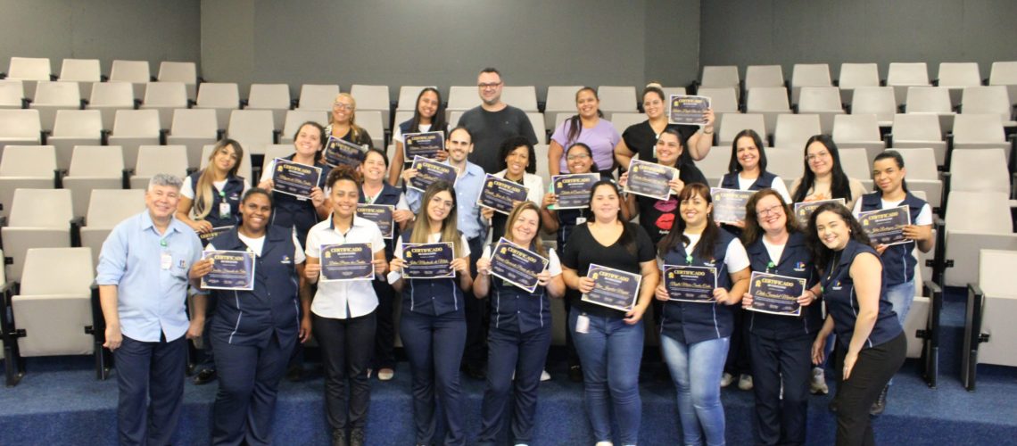 A imagem mostra um grande grupo de pessoas, principalmente mulheres, sentadas em degraus em uma sala de aula. Elas estão segurando certificados e sorrindo para a câmera. Um homem está parado atrás delas. O cenário é uma sala de aula com cadeiras em degraus. A iluminação é boa, e o foco está nas pessoas na frente. Elas parecem estar celebrando uma conquista. A foto sugere uma cerimônia de formatura ou premiação. O ambiente é formal e alegre.