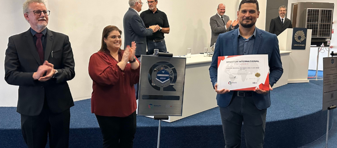 Na imagem, três pessoas se destacam, Nas mãos de um deles, o certificado da QMentum, enquanto os outros interagem com palmas.